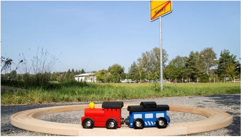 Zwei Holzeisenbahnwaggons stehen auf einer Holsschleife. Im Hintergrund gründende Landschaften, ein Ortsausgangsschild von Niesky ziert den Hintergrund des Bildes.