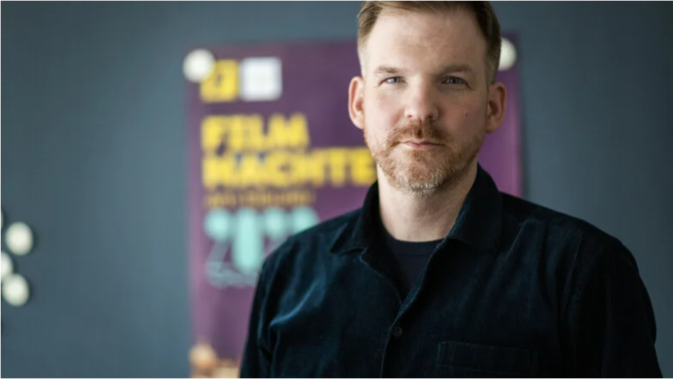 Philip Hartmanis steht im schwarzen Hemd vor einem Plakat der Filmnächte.