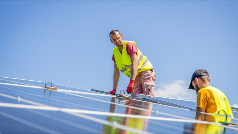 Zwei Männer in gelben Sicherheitswesten stehen auf einem Dach und installieren blaue Solarplatten.