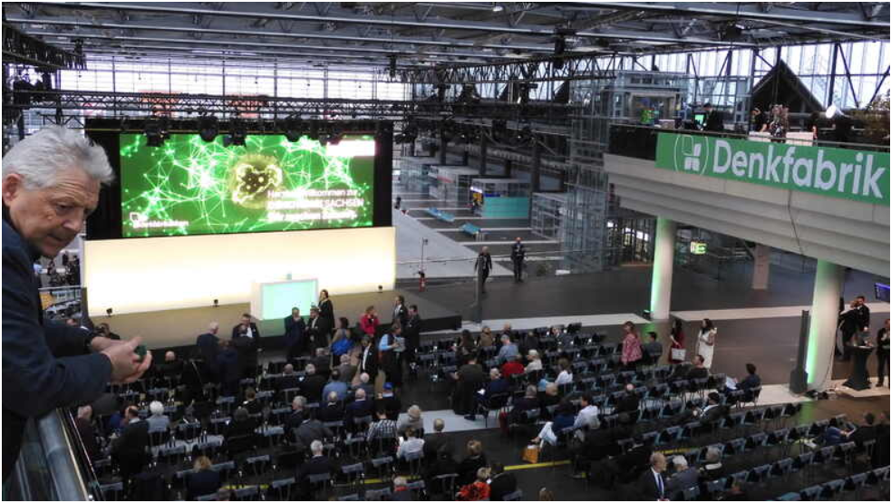 Eine große Leinwand mit Rednerpult steht auf der Veranstaltungsfläche im Dresdner Flughafen. Davor reihen sich viele Stühle mit einigen Gästen der Tagung.