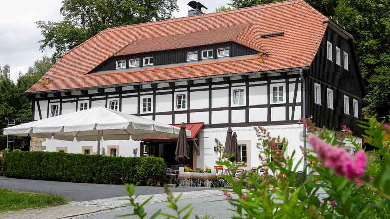 Der Gasthof "Alte Schmiede" in Lückendorf ist ein altes, großes Fachwerkhaus mit rotem Satteldach. Vor dem weiß gestrichenen Haus ist ein cremefarbener Sonnenschirm aufgespannt, unter dessen Plane sich Terassenmöbel befinden. Eine grüne, hüfthohe Hecke zäunt das Gelände ein.