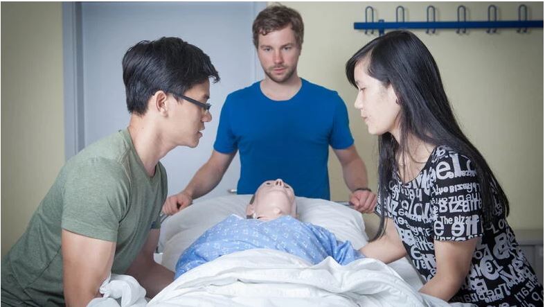 Drei Auszubildende stehen um eine Puppe im Krankenbett und üben für Ihre pflegerische Ausbildung.