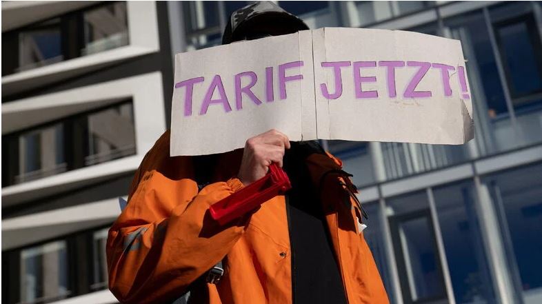 Ein Tarifstreiker steht mit einem Schild mit der Aufschrift "Tarif jetzt" vor dem Gesicht vor einem Haus.