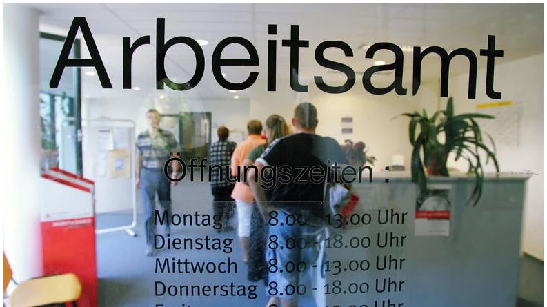Man sieht die Aufschrift "Arbeitsamt" auf einer Glastür, hinter welcher Menschen in einer Schlange am Schalter warten.