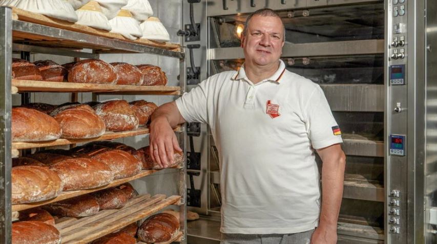 Man sieht Bäckermeister Roland Ermer in seiner Bäckerei mit vielen frisch gebackenen Broten.