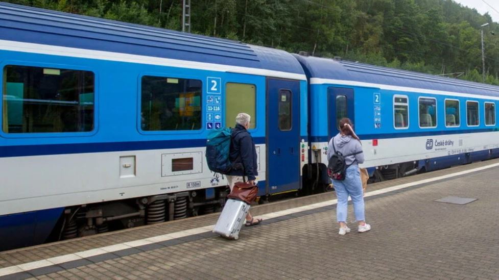 Man sieht 2 Passagieren vor einem Zug mit ihren Koffern und Taschen.