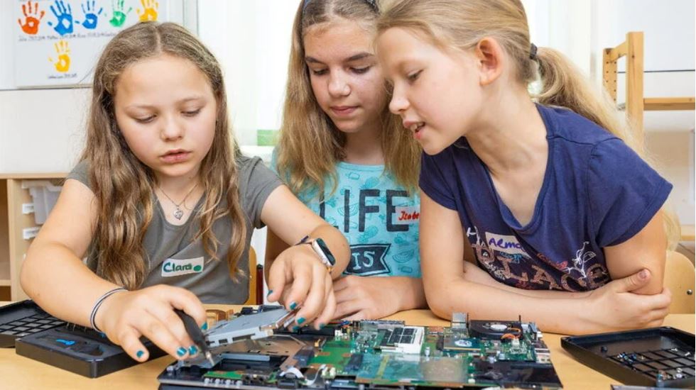 Man sieht drei Schülerinnen, die gerade lernen wie man einen Laptop auseinander baut