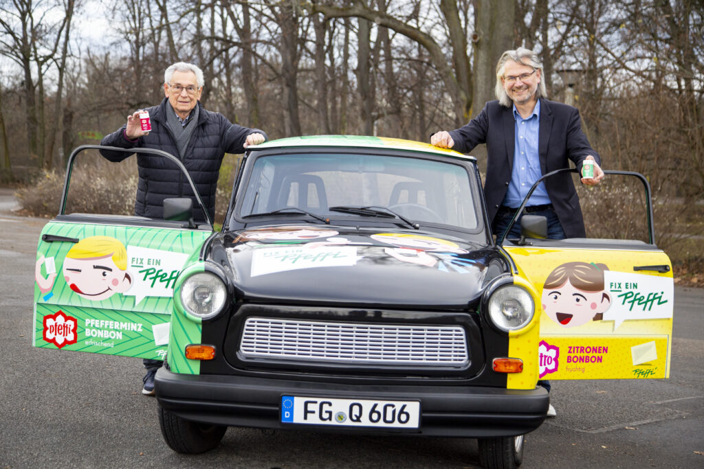 zwei Männer stehen lächelnd neben einem Trabant, der mit Werbung für "Pfeffi" bedruckt ist.