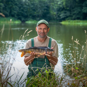 Ein Mann steht am See und hält einen Karpfen in der Hand.