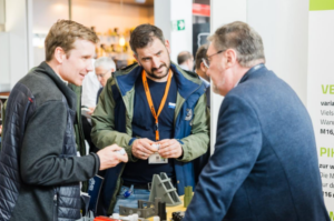 Drei Männer stehen an einem Messestand und begutachten gemeinsam Kunststoffbauteile. Im Hintergrund weitere Messegäste und ein Ausstellerbanner.