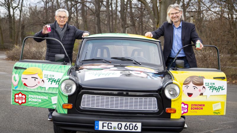 zwei Männer stehen lächelnd neben einem Trabant, der mit Werbung für "Pfeffi" bedruckt ist.